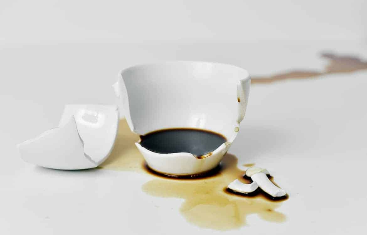 a broken white ceramic cup of coffee with its pieces and the coffee spilled on a white table